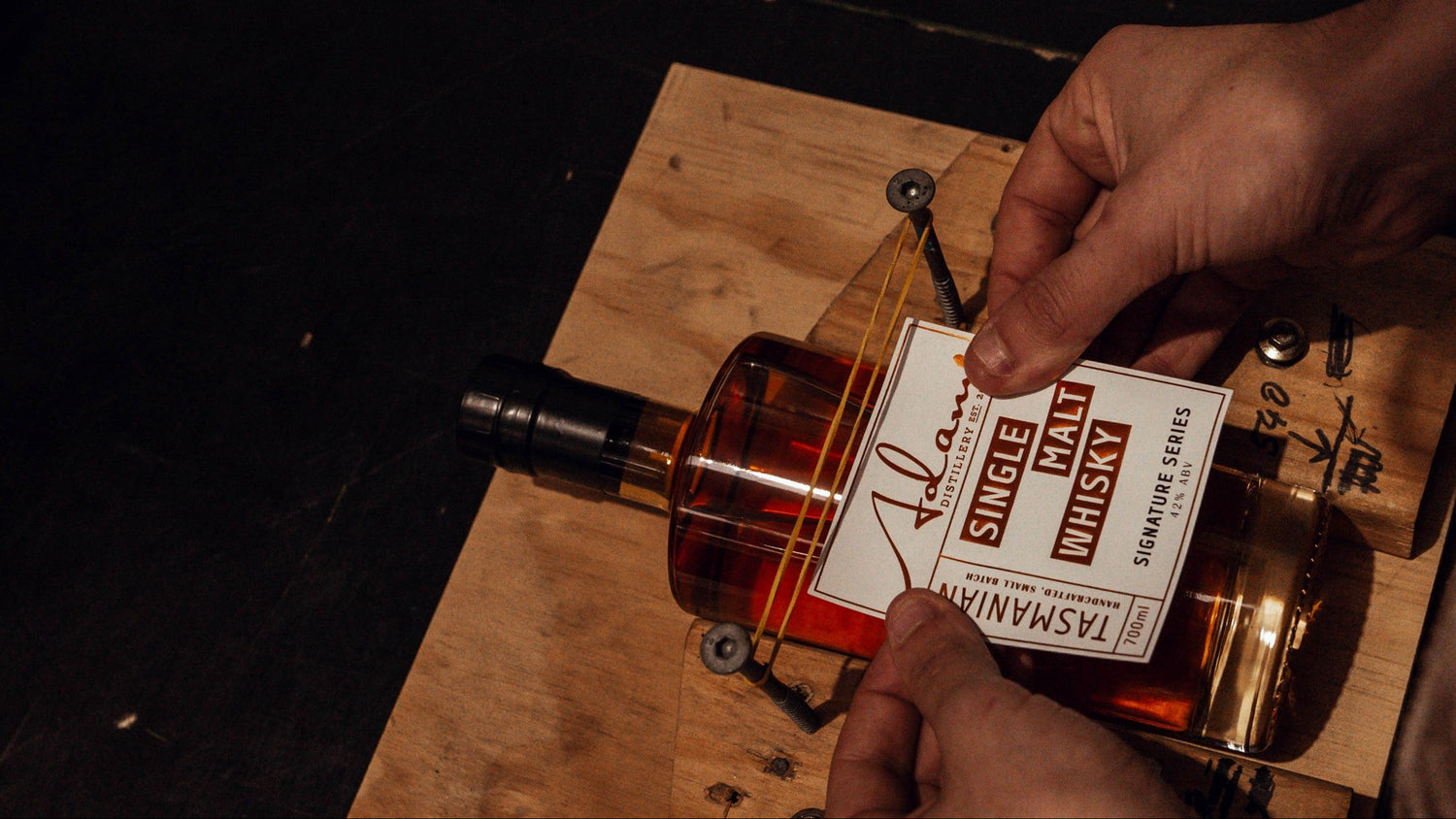 Person holding an Adams bottle of 'Single Malt Whisky' with a wooden label on a wooden surface.